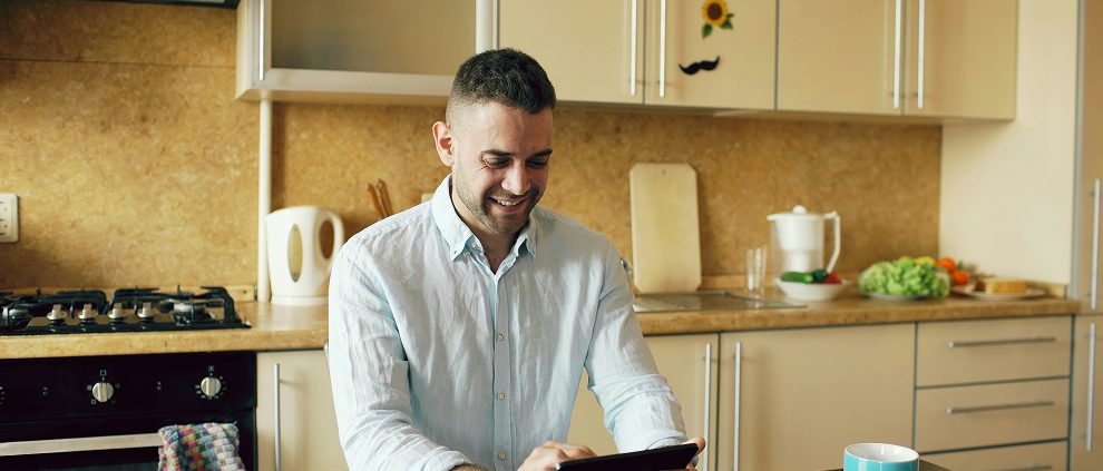 Smiling man in a light blue shirt uses a tablet in a bright kitchen with light brown cabinets.