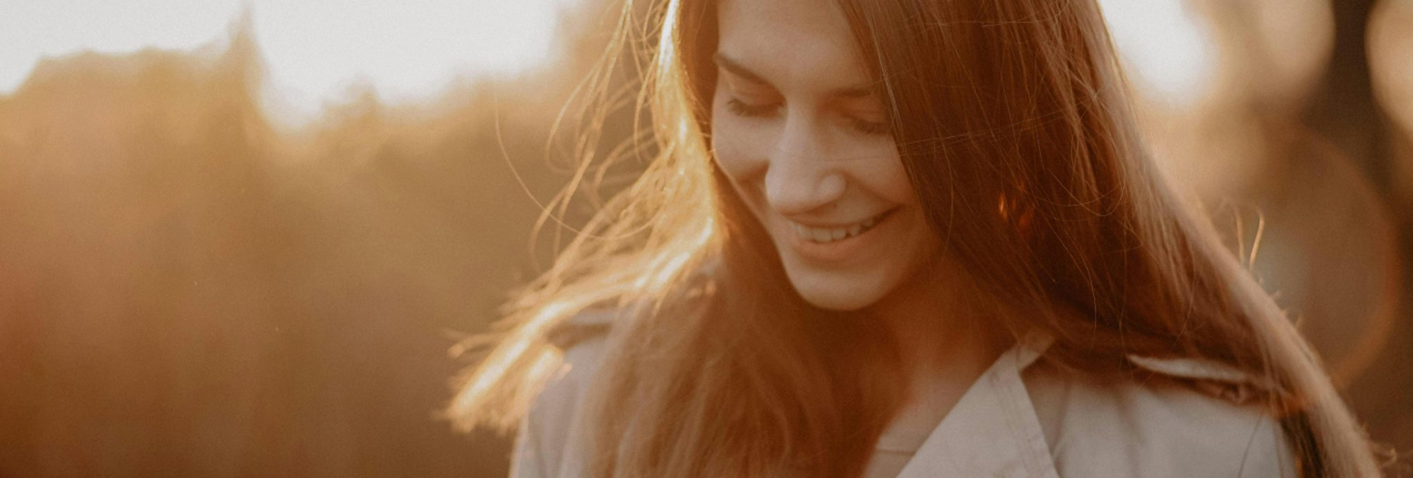 A woman with long, flowing hair smiling softly, illuminated by warm, golden sunlight.
