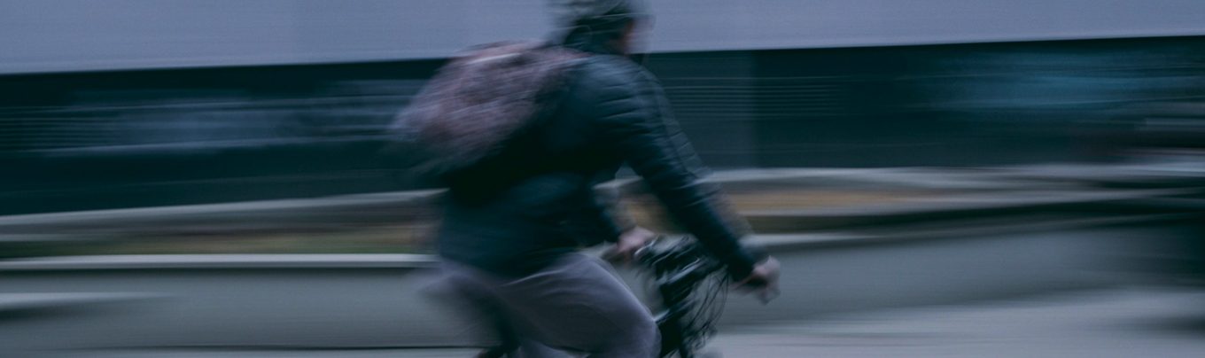 Motion blur shot of a person cycling, wearing a jacket and backpack, against a streaky background.