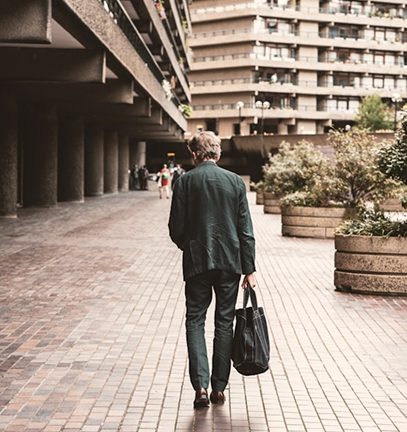 Man in a suit walks away with a bag through a brutalist urban complex with concrete buildings.