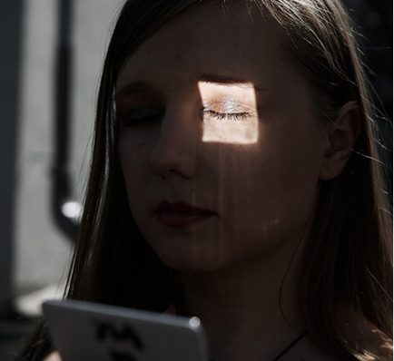 Woman with closed eyes, a square of sunlight on her left eye. She holds a phone.