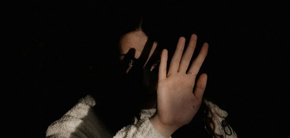 A person's hand covers part of their face in shadow, with one eye visible and a ring on a finger.