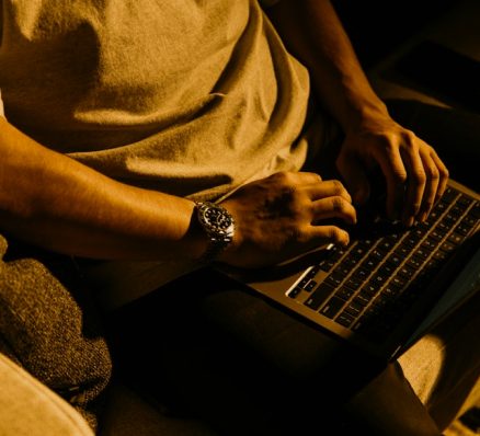 Person's hands typing on a laptop keyboard in a dimly lit setting, wearing a watch.