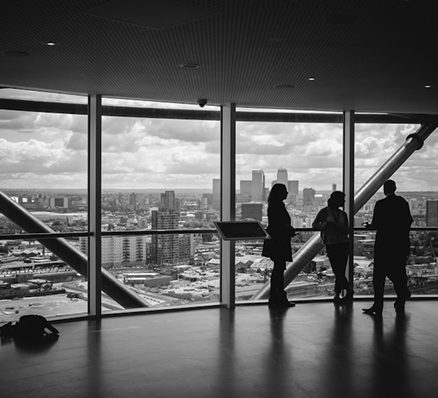 Black and white image of three silhouettes looking out large windows at a city skyline.