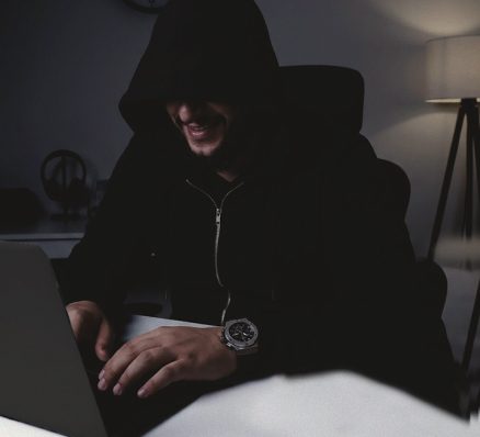 A person in a black hoodie laughs while typing on a laptop in a dimly lit room.