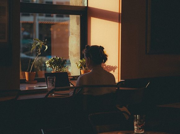 A person sits with their back to the viewer, working on a laptop by a window with warm sunlight.