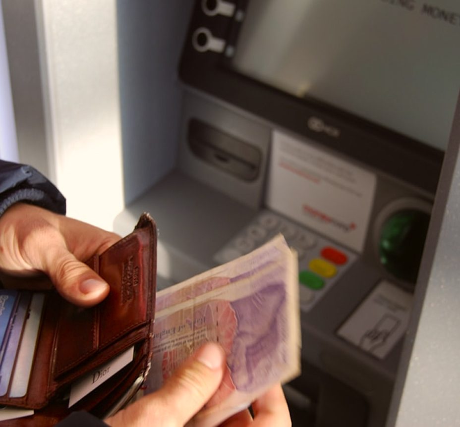 A person is holding a brown leather wallet and British pound banknotes while standing in front of an ATM.