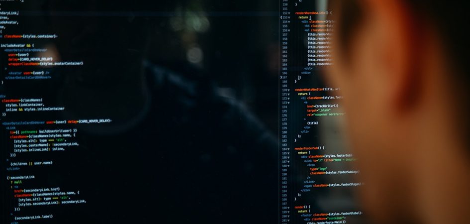 Close-up of a person's blurred head watching code on a dark computer screen, lines of green and white text visible.