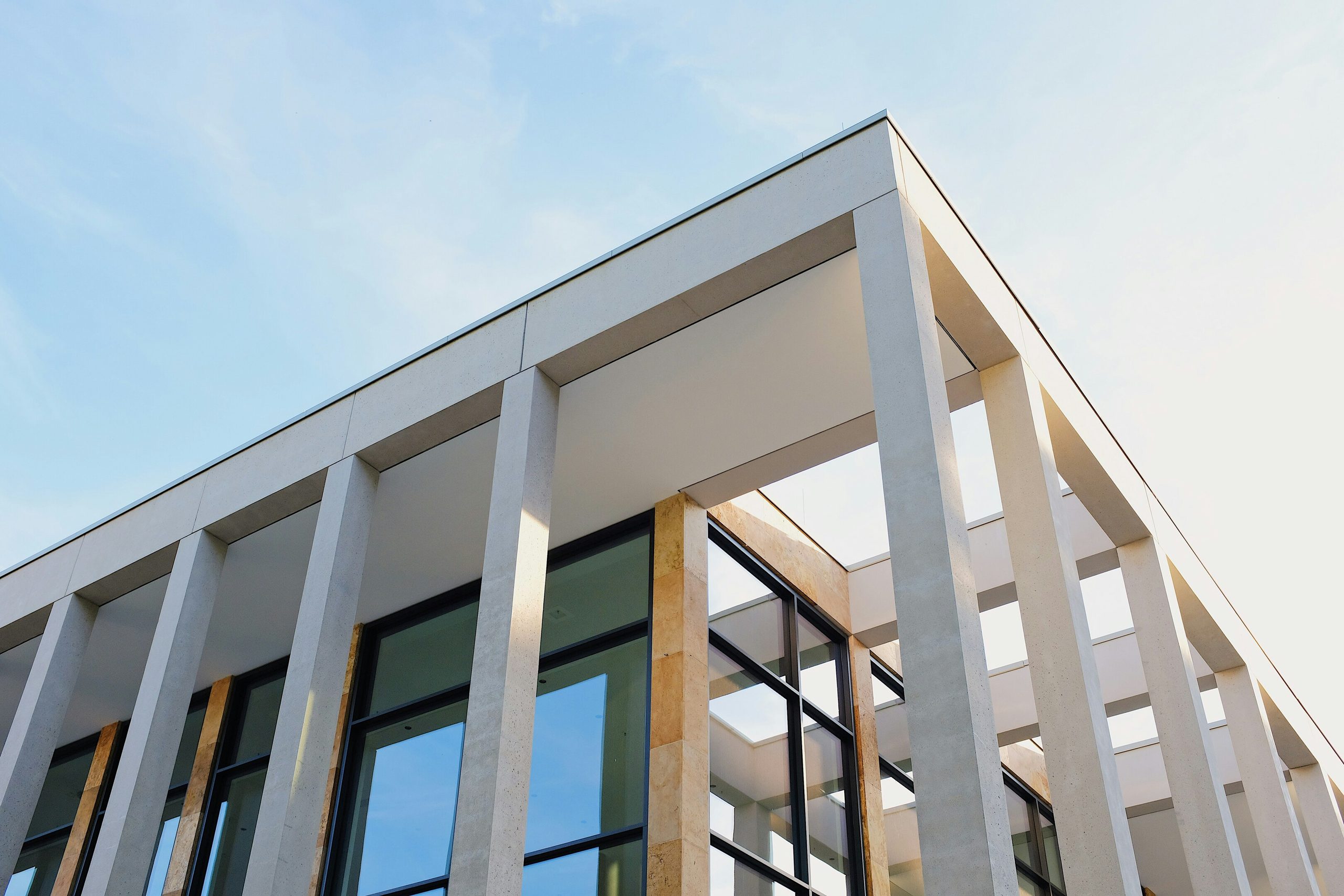Modern building facade with light concrete columns and reflective glass windows against a blue sky.