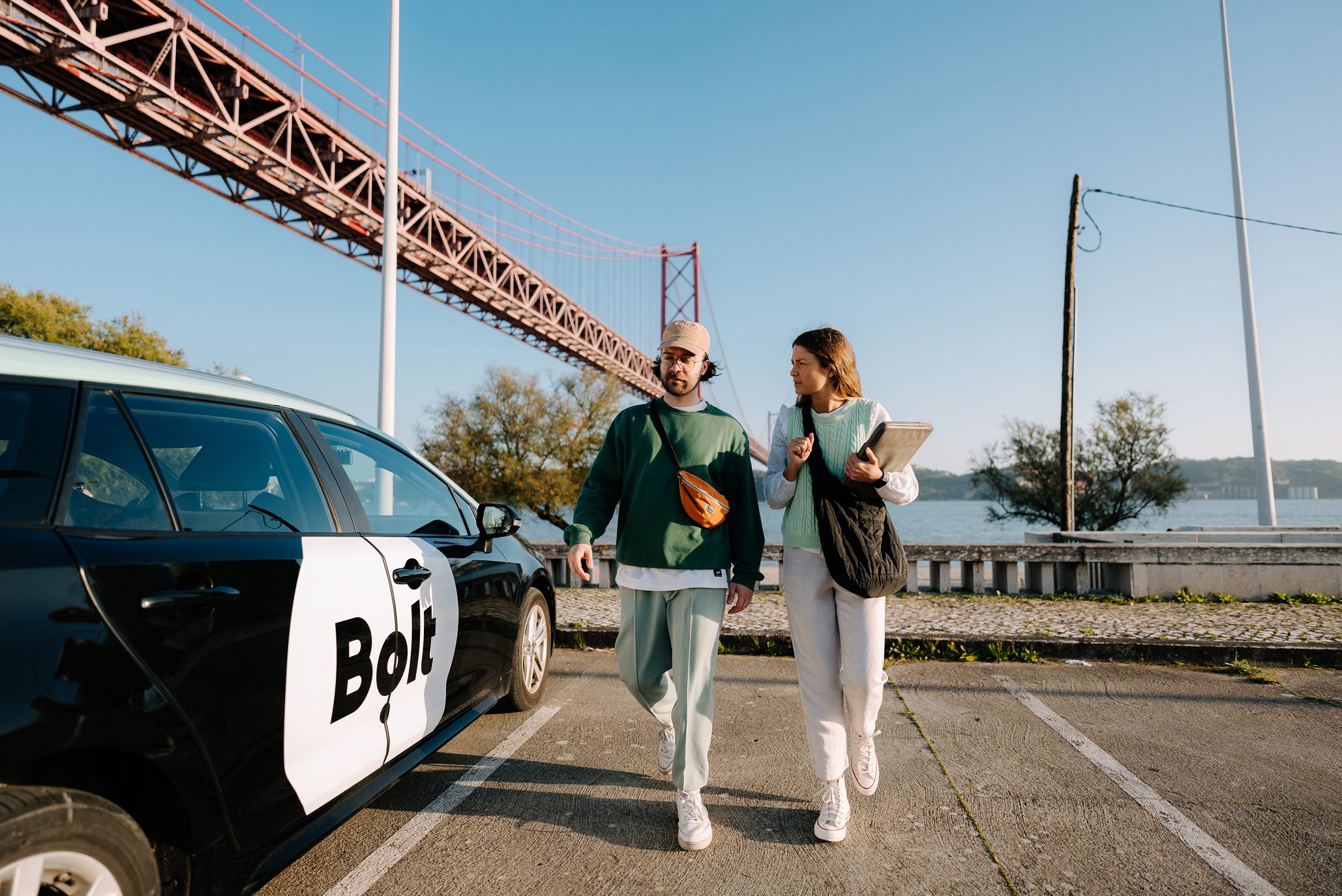 Two people walking past a Bolt ride-sharing car with a large suspension bridge in the background on a sunny day.