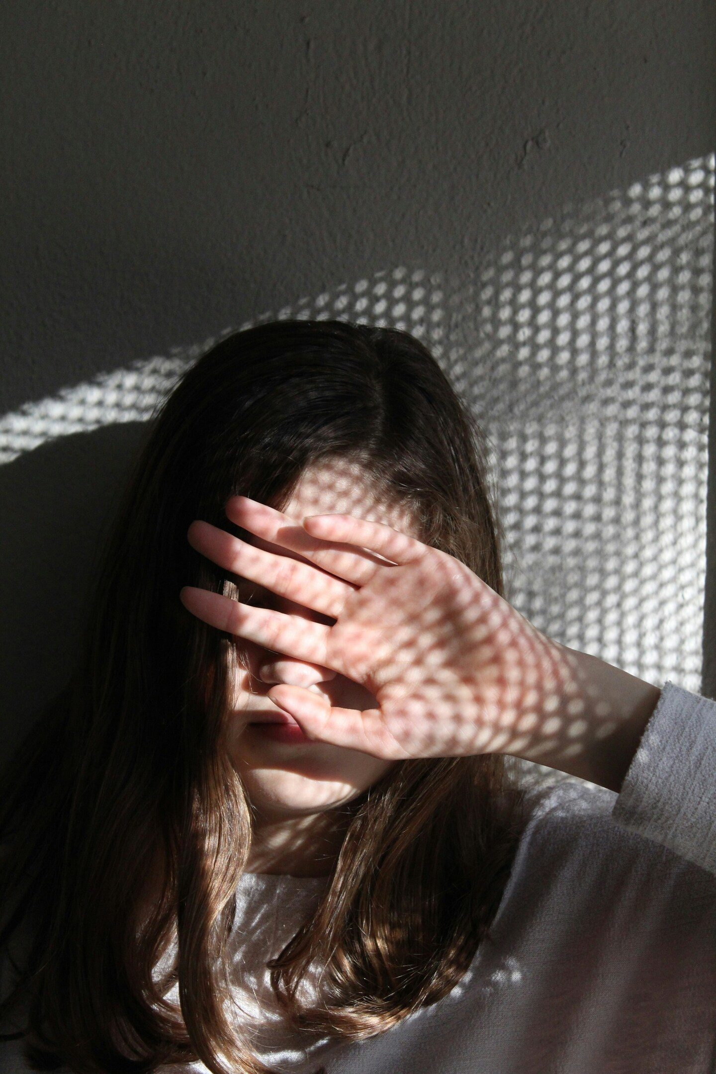 A person's face covered by a hand, with dappled sunlight creating a grid pattern over their skin and hair.