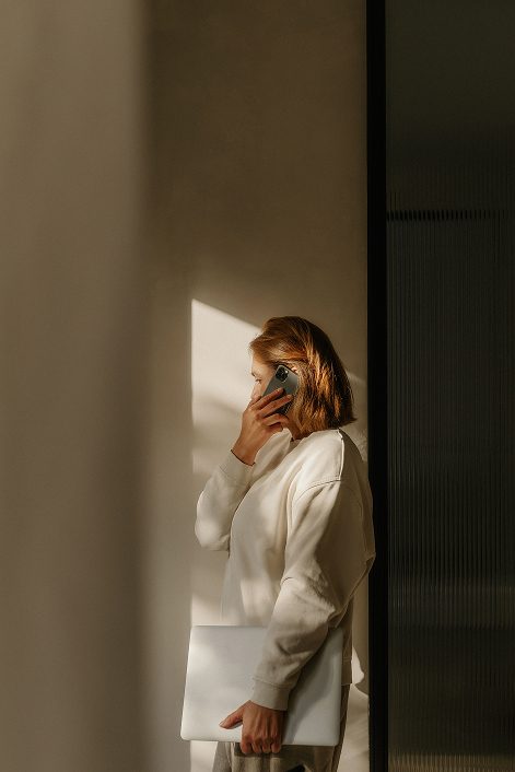 Woman on phone holding a laptop, standing in a sunlit room.