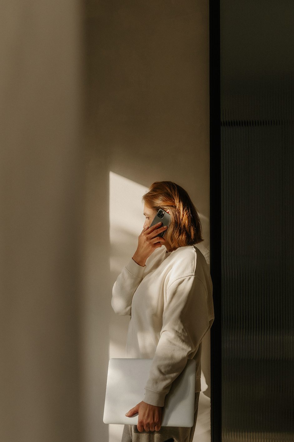 A woman in a sunlit room talking on a phone while holding a laptop, seen in profile.