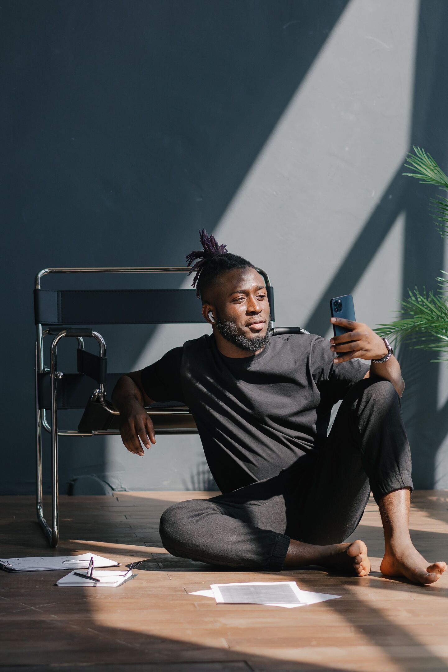 A man with dreadlocks and earbuds sits on the floor, leaning on a chair, holding a phone.