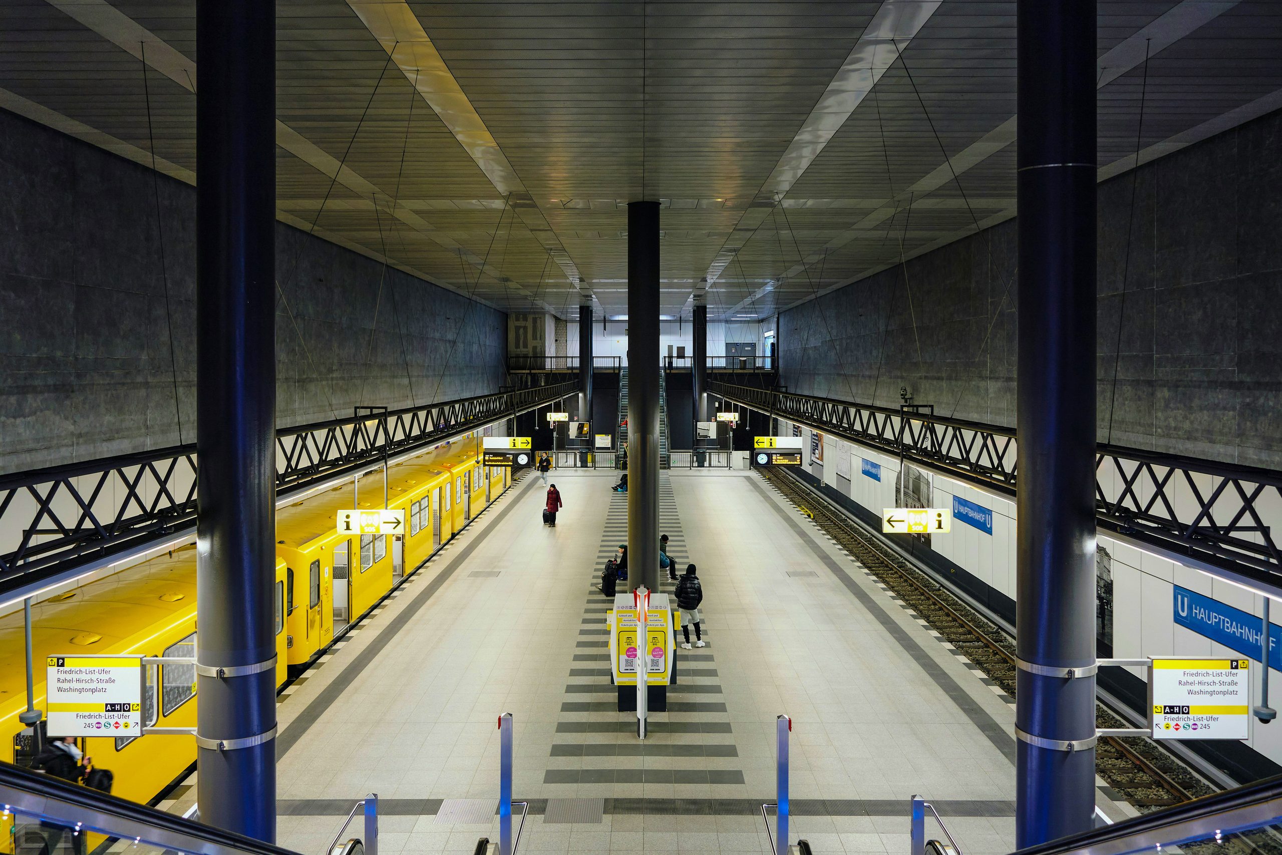 Modern, symmetrical subway station with a yellow train, people on the platform, and a long tunnel.