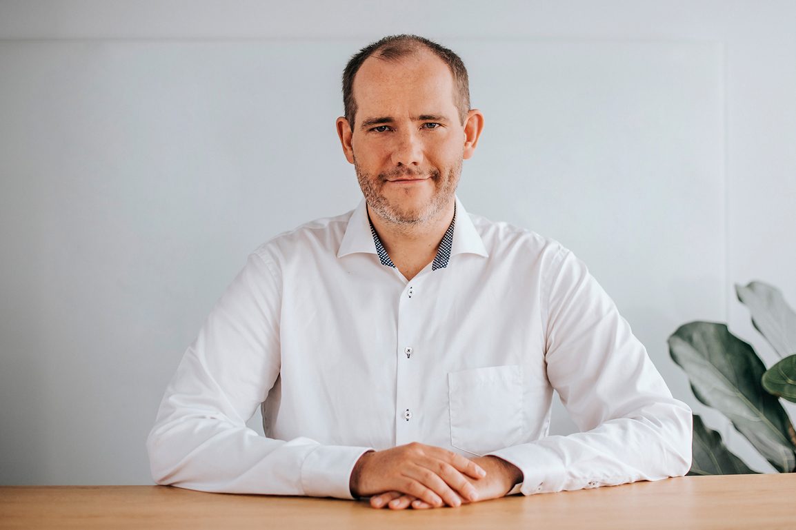 A man with a beard and white shirt is sitting at a table, looking forward.