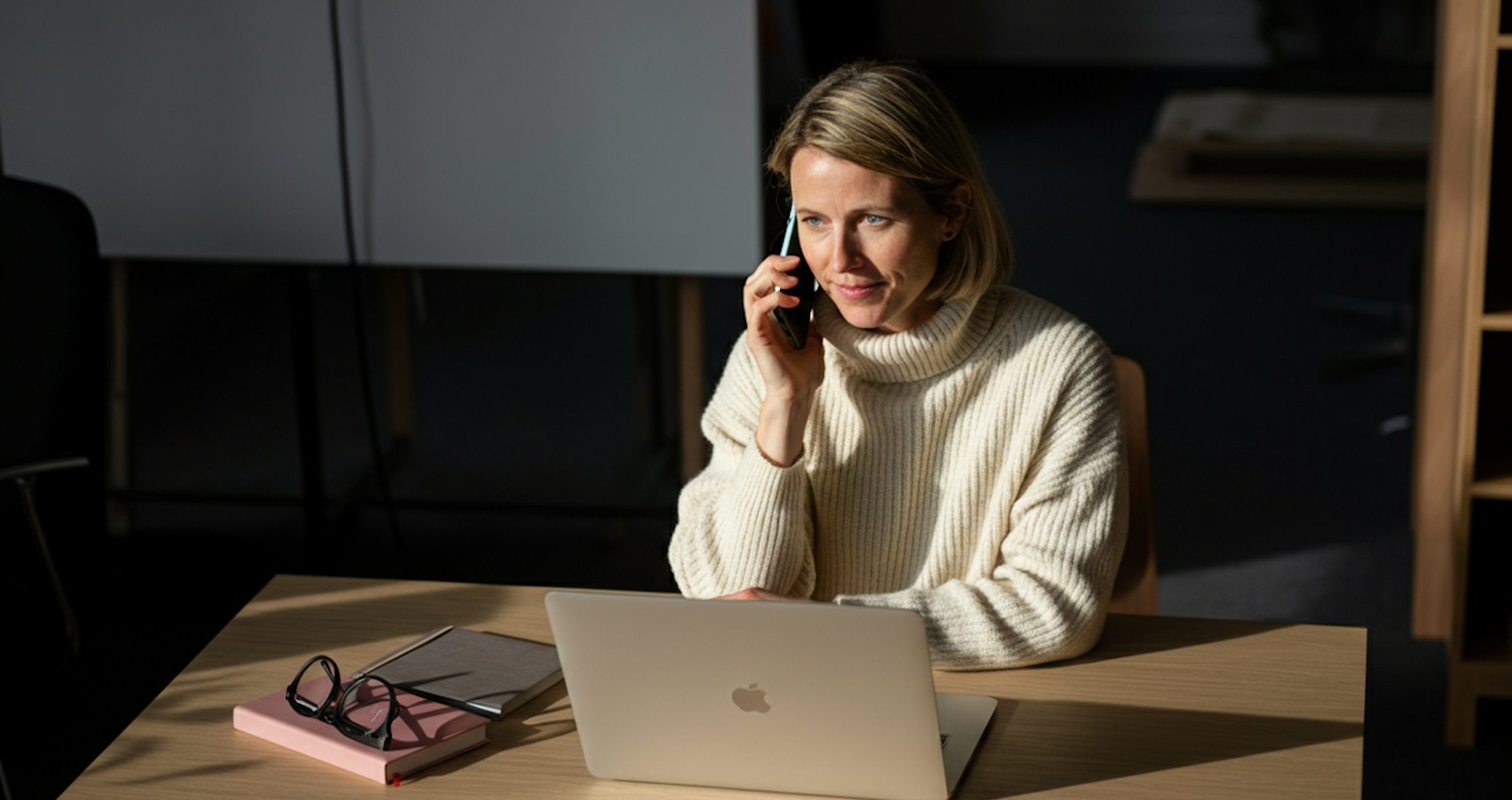 A woman in a cream sweater talks on the phone while looking at a laptop on a wooden desk.