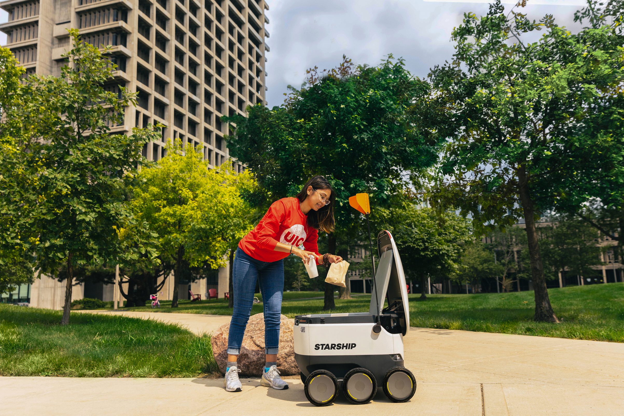 Woman in UIC shirt loading a cup and bag into a Starship delivery robot on a campus path.