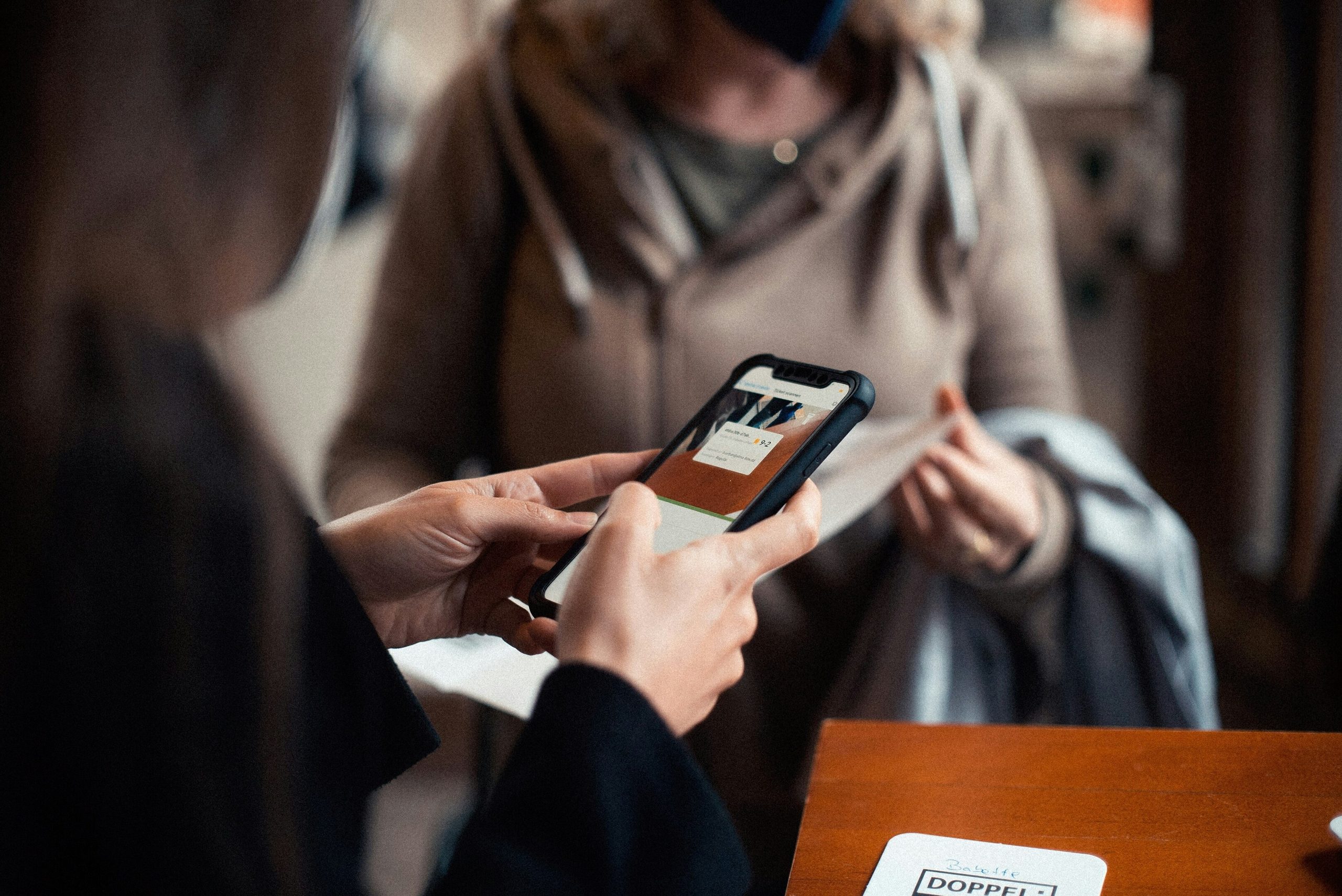 Hands holding a smartphone displaying an interface, with a person in the background.