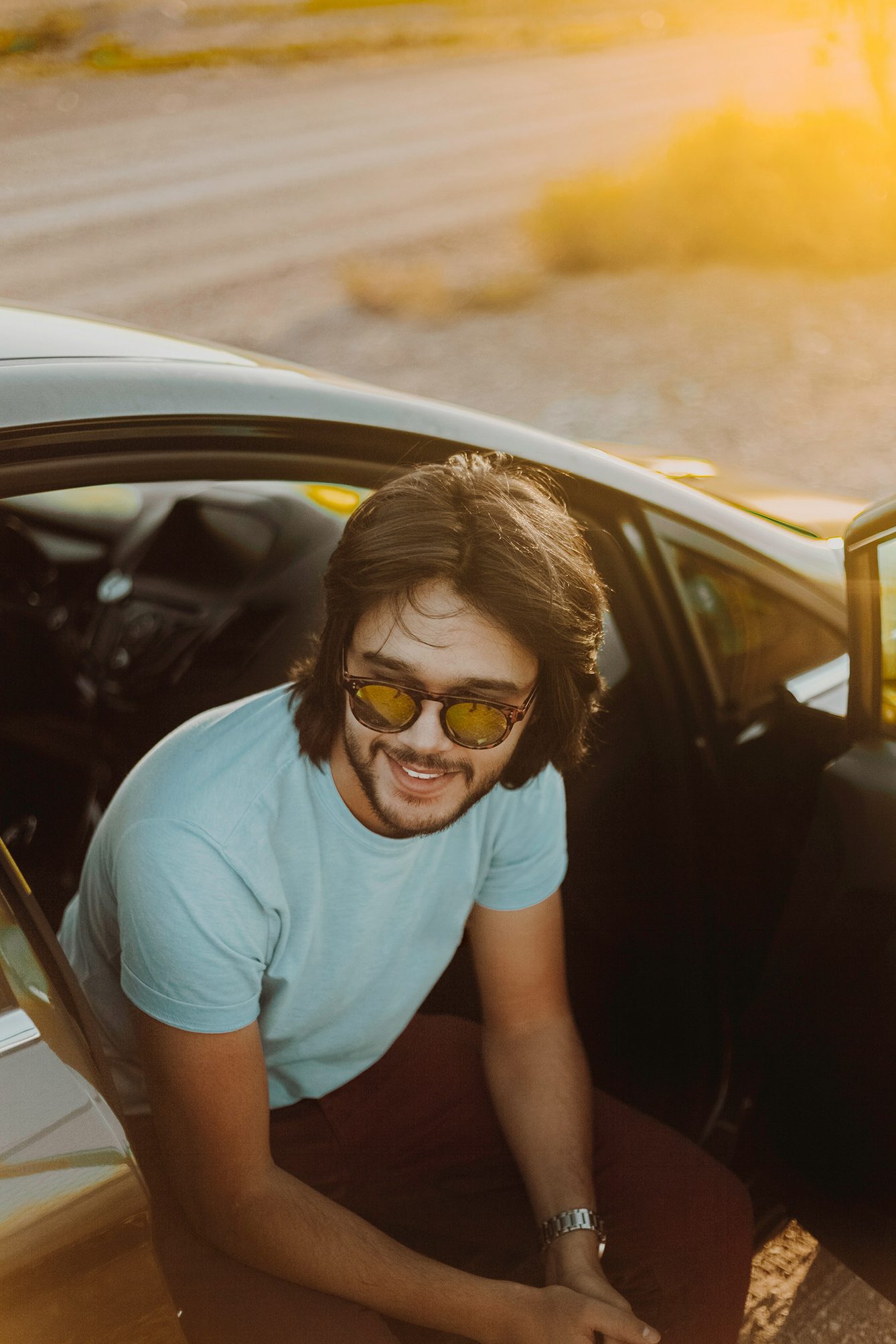 A smiling man with long hair and sunglasses sits in a car doorway during golden hour.