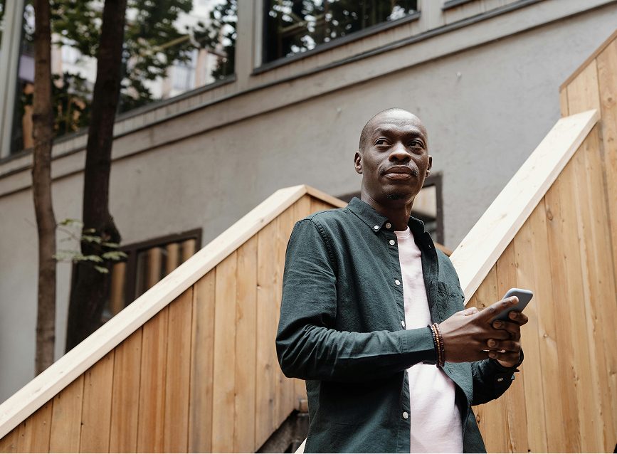 A man with a shaved head and a green shirt looking up while holding a smartphone outdoors.