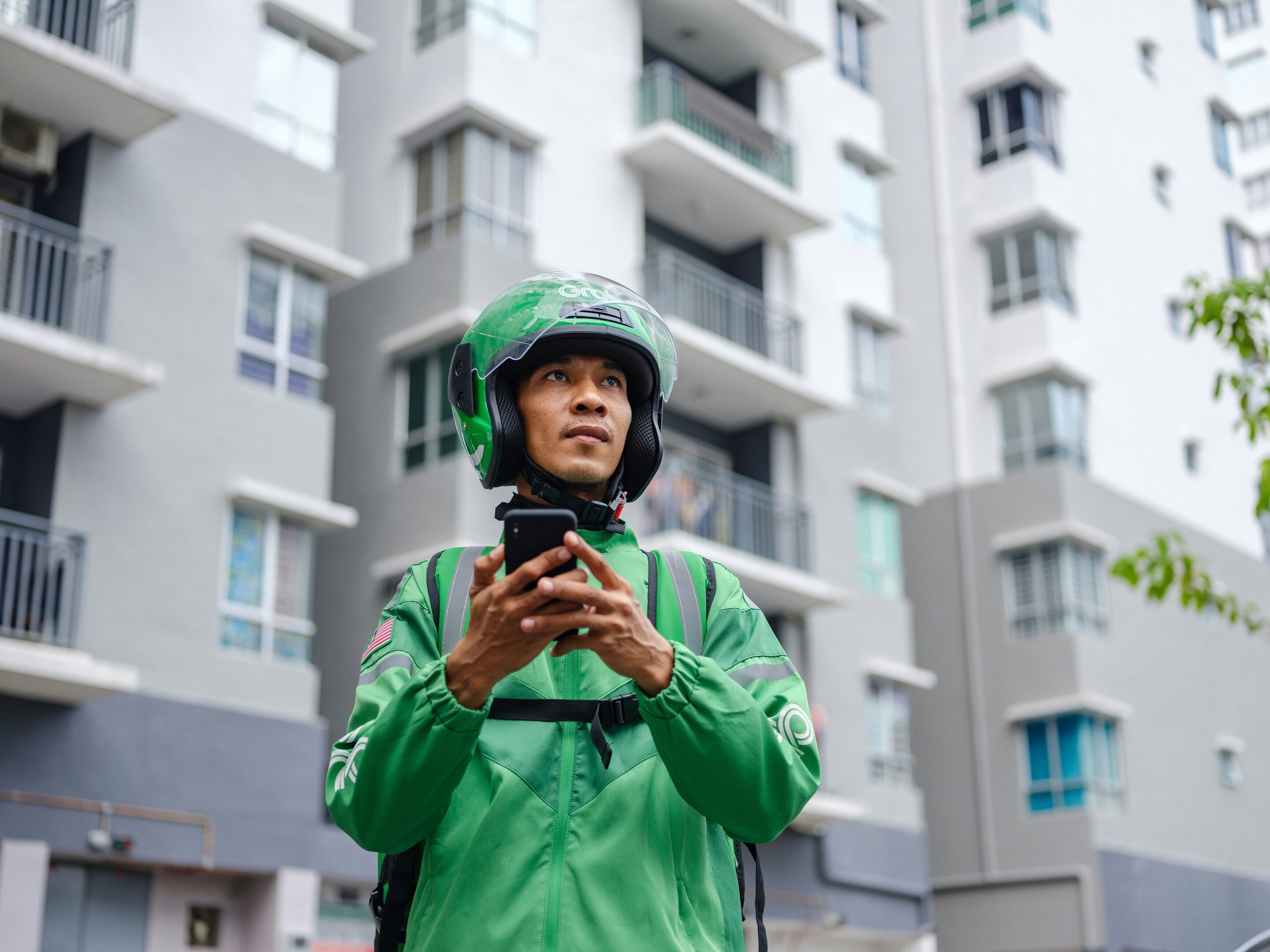 A man in a green delivery uniform and helmet looks up while holding a smartphone against an apartment building.