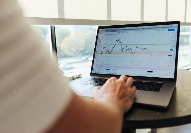 Person's hand typing on a laptop keyboard, with the screen displaying financial charts and graphs, by a window.