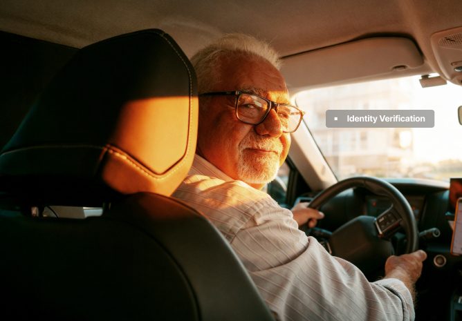 An older man with glasses and a white beard smiles from the driver's seat of a car.