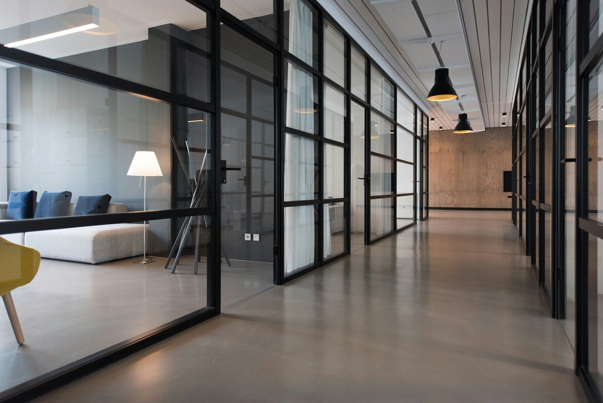 A modern office hallway featuring polished concrete floors, black-framed glass walls, and a lounge area.