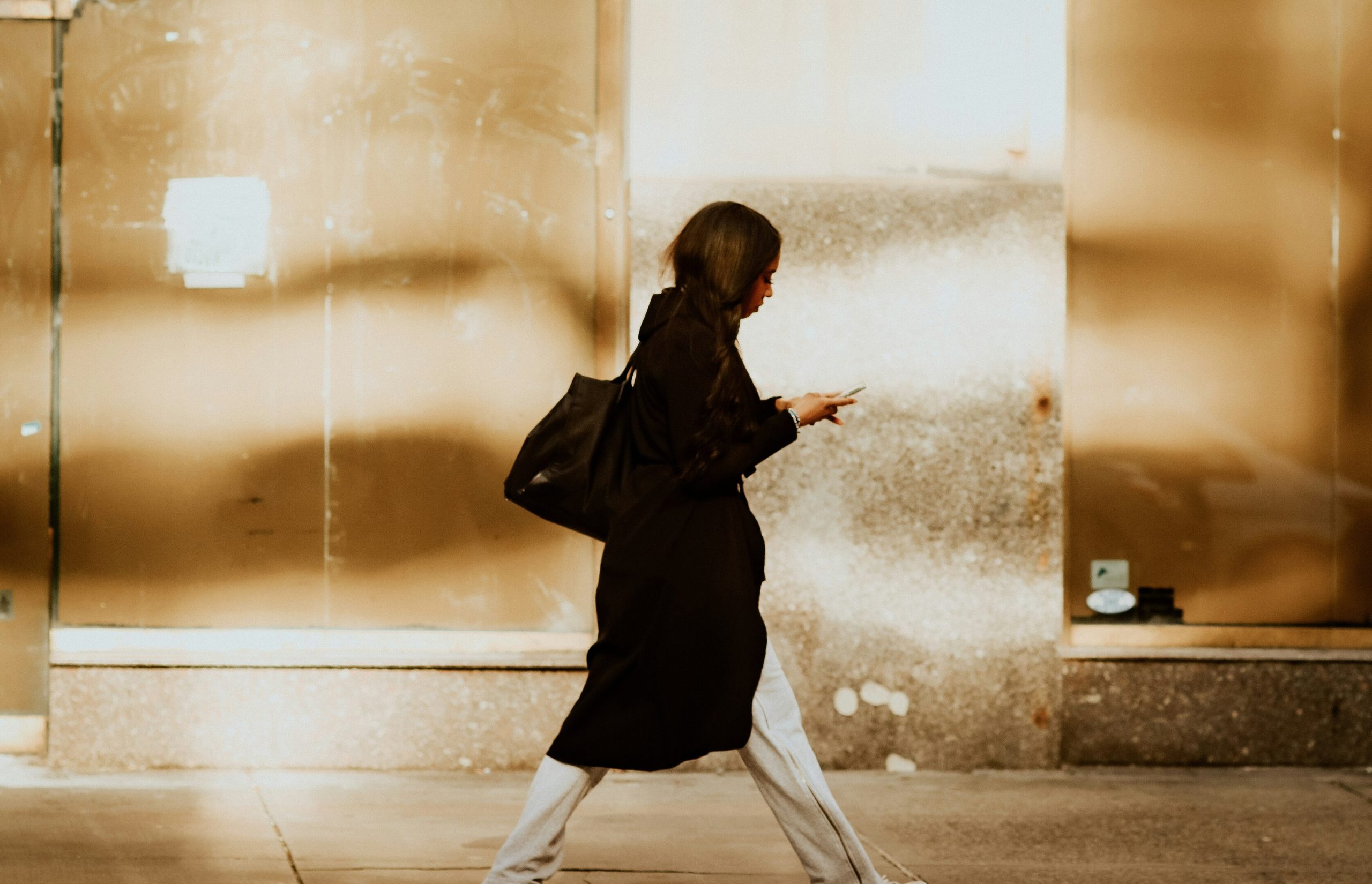 Woman in black coat walks on sidewalk, engrossed in her phone.