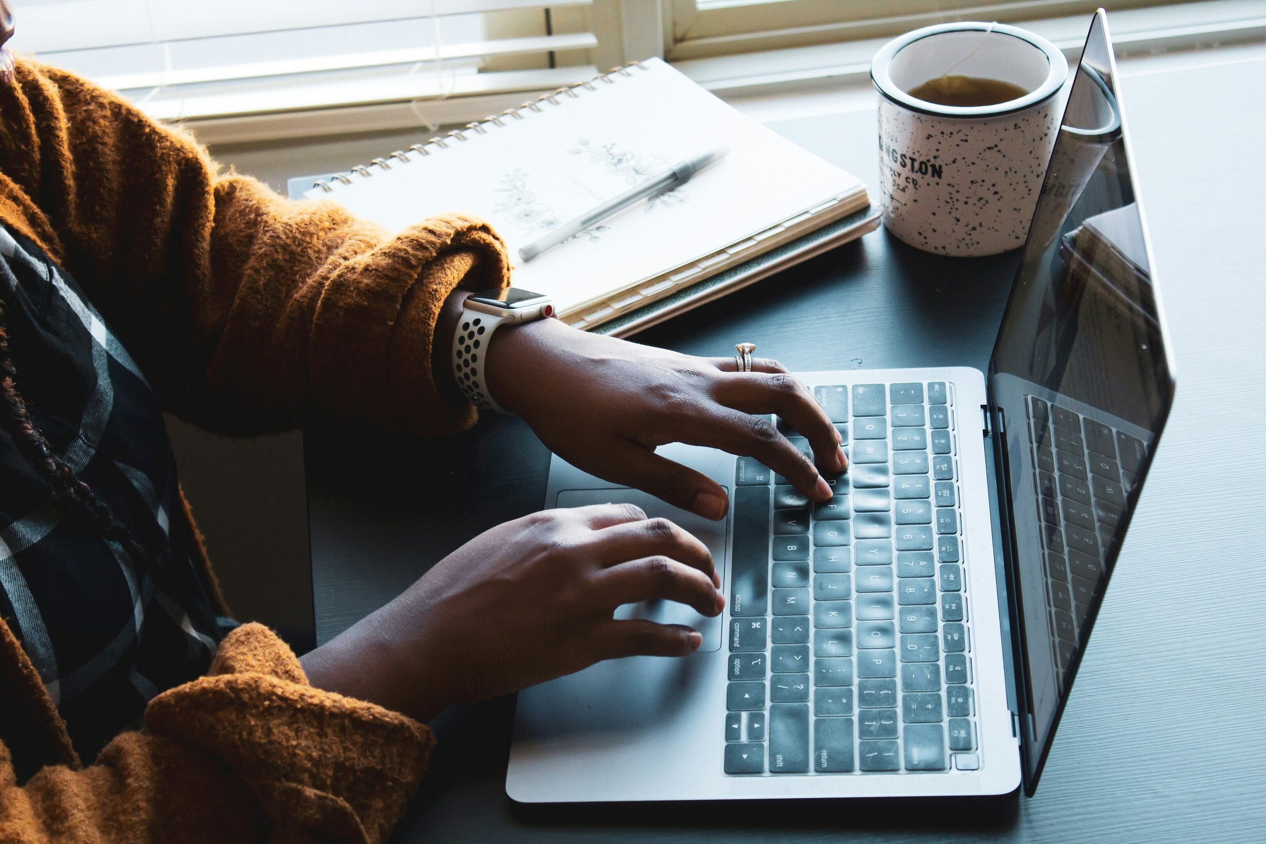 A person's hands typing on a laptop keyboard, with a notebook, pen, and coffee mug on a desk.