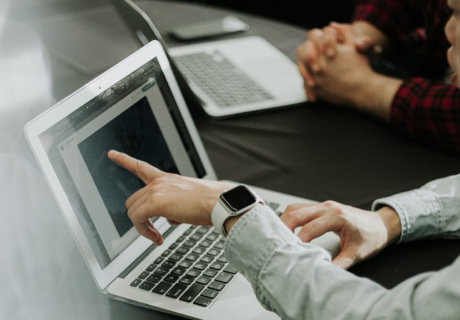 A person's hand with a smartwatch pointing at a laptop screen during a collaborative meeting.