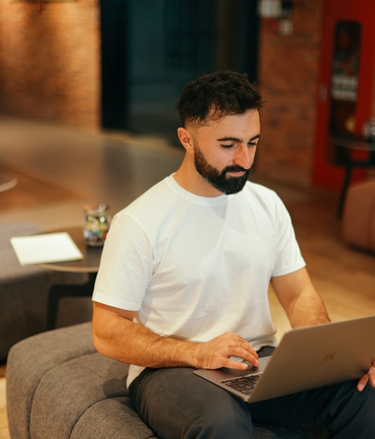 Man with beard and white t-shirt using a laptop, sitting on a gray cushion.