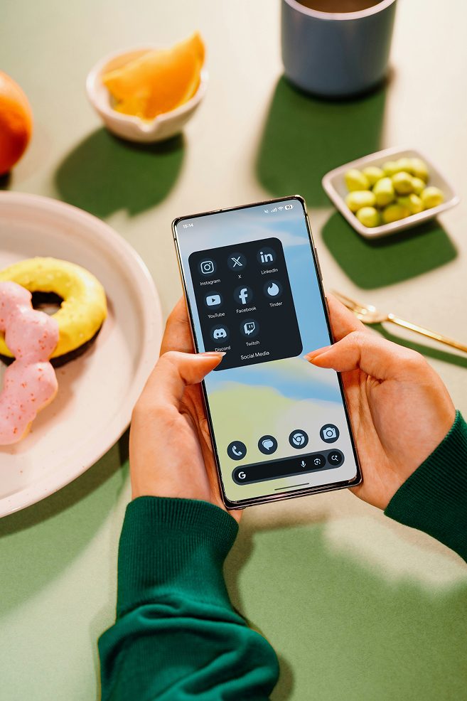 Hands holding a smartphone displaying a social media folder, with snacks on a table.